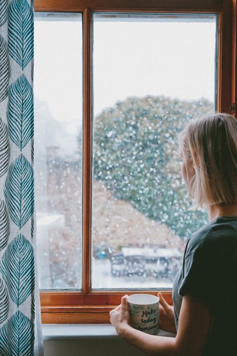 nő csészével kifelé néz a meleg otthon ablakán a hóesésébe